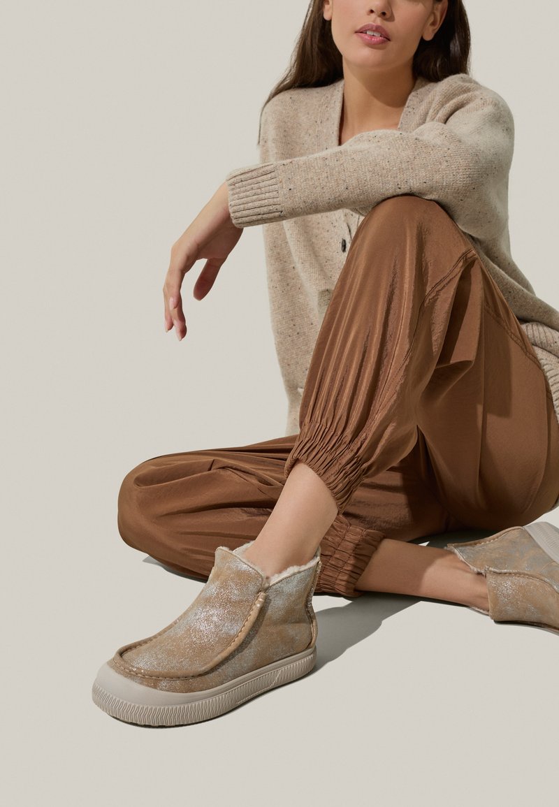Scarpe beige slip-on con una brillantezza metallica, fodera morbida e suola in gomma texturizzata. Abbinato a pantaloni marroni con fondo arricciato e un maglione leggero.