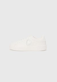 Λευκό παπούτσι τύπου sneaker χαμηλού ύψους με κορδόνια, στρογγυλεμένη μύτη και λογότυπο σε σχήμα ασπίδας στη πλαϊνή πλευρά, τοποθετημένο σε απλό φόντο.