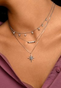 Colliers en argent superposés présentant des motifs d'étoiles, un pendentif en barre texturée et un charm en forme d'étoile central, sur un fond de teint et de tissu.