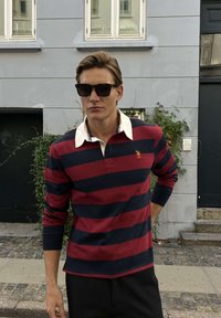 Férfi fekete napszemüveget visel, piros és tengerészkék csíkos rugbypólót fehér gallérral, szürke fal előtt állva a szabadban.