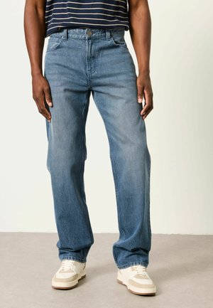 Person trägt blaue Jeans mit geradem Bein, weiße Turnschuhe und ein blau-weiß gestreiftes Hemd und steht auf einem grauen Boden.