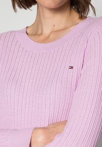 Maglione lavorato a maglia di colore rosa chiaro con scollo rotondo, caratterizzato da una texture a coste e un piccolo logo accento sul petto. Dettagli orizzontali sottili.