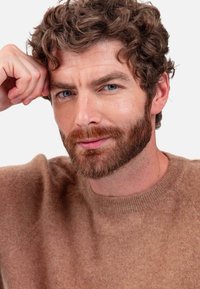 Light brown wool sweater with a crew neck and textured finish. The model has curly hair and a short beard, resting his chin on his hand.