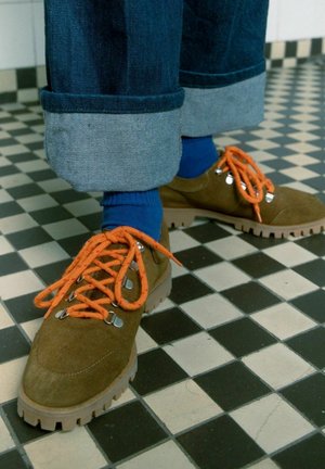 Bruine suède schoenen met oranje veters, metalen oogjes en gestructureerde zolen. Draag ze met opgerolde blauwe denim jeans en blauwe sokken op een betegelde vloer.