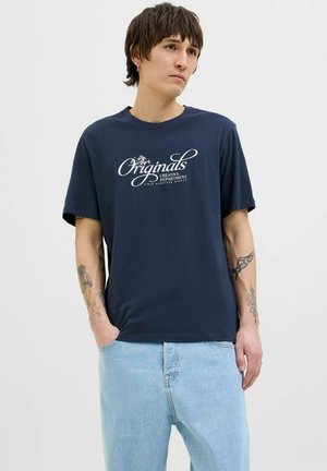 Donkerblauwe katoenen t-shirt met korte mouwen, met een witte grafische tekst "Originals Creative Department" op de voorkant. Lichtblauwe spijkerbroek.