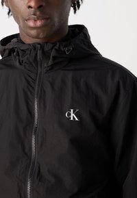 Črna lahka jakna s kapuco, ki ima sprednji zadrgo in majhen bel "ck" logo na levem delu prsnega koša. Smoother, mat tekstura.