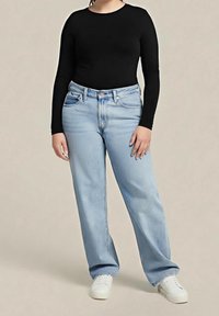 Personne portant un haut noir ajusté à manches longues, un jean large taille haute bleu clair et des baskets blanches, debout devant un fond beige.