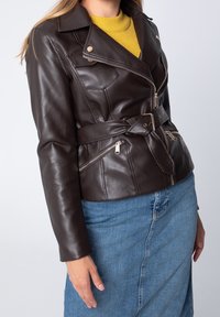 Ruskea tekonahkainen takki, jossa on vyötäröllä vyö, vetoketjulliset taskut ja metalliset yksityiskohdat; yhdistetty keltaiseen neulepaitaan ja farkkuhameeseen.