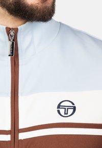 Trainingsjack met een lichtblauwe bovensectie, witte middelste sectie met bruine strepen en een logo. Ritsdetails bij de kraag en een gestructureerde stof.
