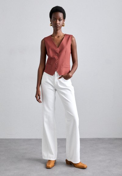 Chaleco sin mangas de color óxido con escote en V, cinco botones y dos bolsillos frontales. Combinado con pantalones anchos blancos y zapatos color marrón claro.
