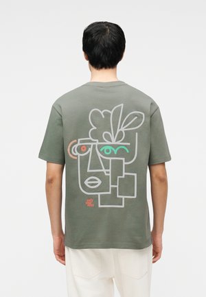 Homme portant un t-shirt vert olive avec un dessin abstrait de visage et de feuille en lignes blanches, rouges et bleu sarcelle au dos, associé à un pantalon blanc.
