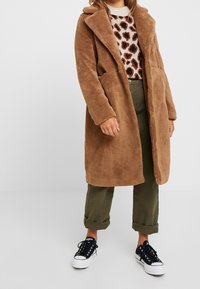 Persona vestida con un abrigo largo marrón de felpa, un suéter de estampado de leopardo, pantalones verde oliva remangados y zapatillas en blanco y negro.