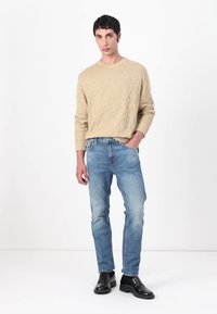 Beige tekstuuriga sviiter ümmarguse kaelusega, koos heledate siniste teksadega. Mustad kingad. Mudel seisab ühtlase valge tausta ees.