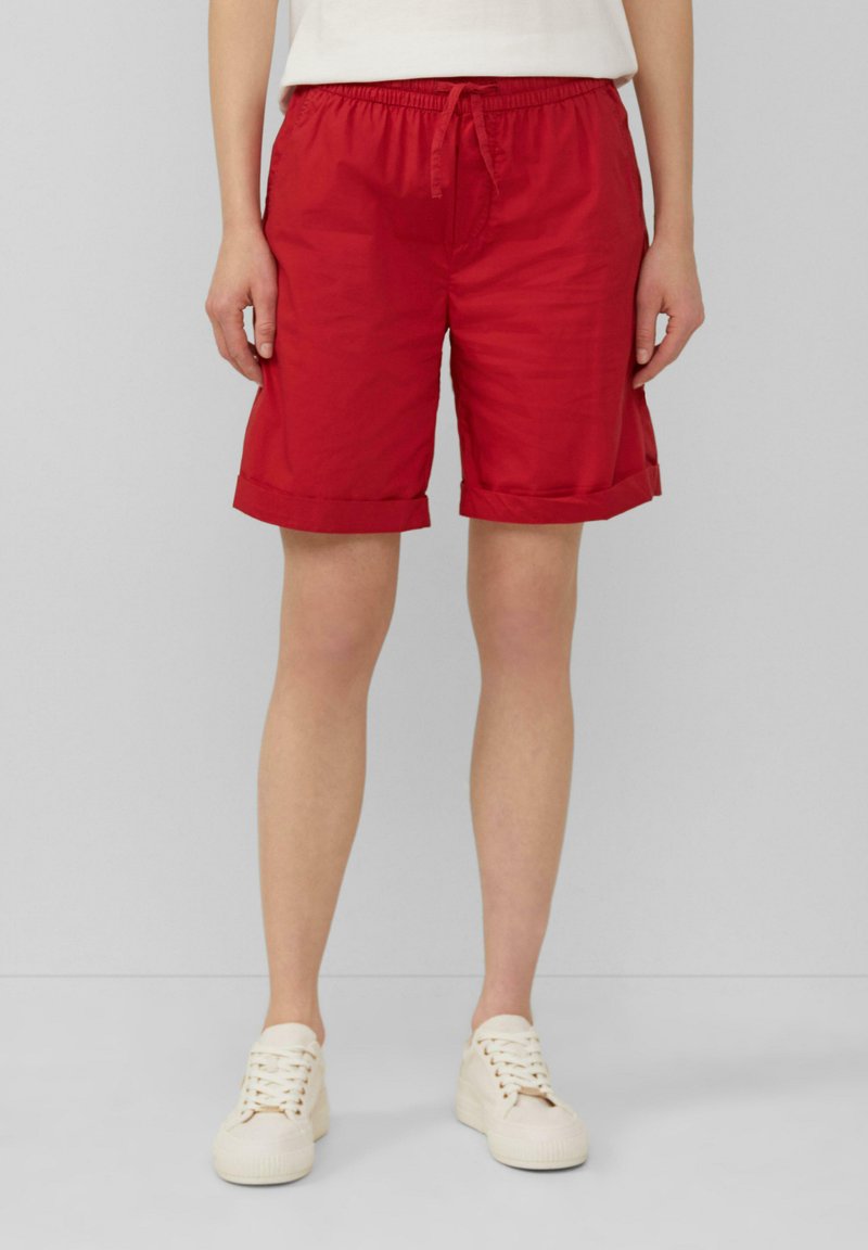 Rode shorts met een elastische tailleband en trekkoord, met opgerolde mouwen en een gladde, licht glanzende textuur. Draagt een lichtgekleurde sneakers.