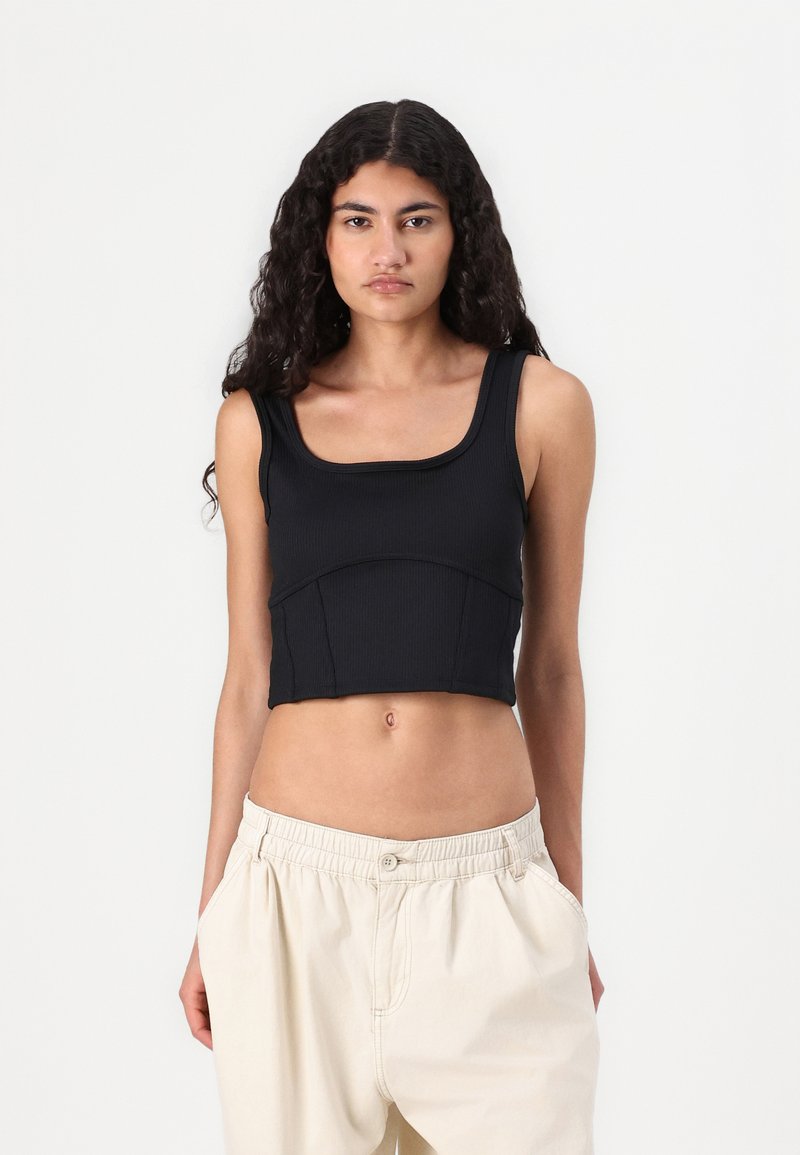 Črn rebrast crop top s kvadratnim izrezom, oprijet kroj in šivi na pasu, v kombinaciji z bež ohlapnimi hlačami.