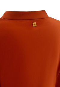 Orange Bluse mit glatter Textur, ausgestattet mit einem Kragen und einem kleinen goldenen Tag auf der Rückseite, der den Markennamen zeigt.