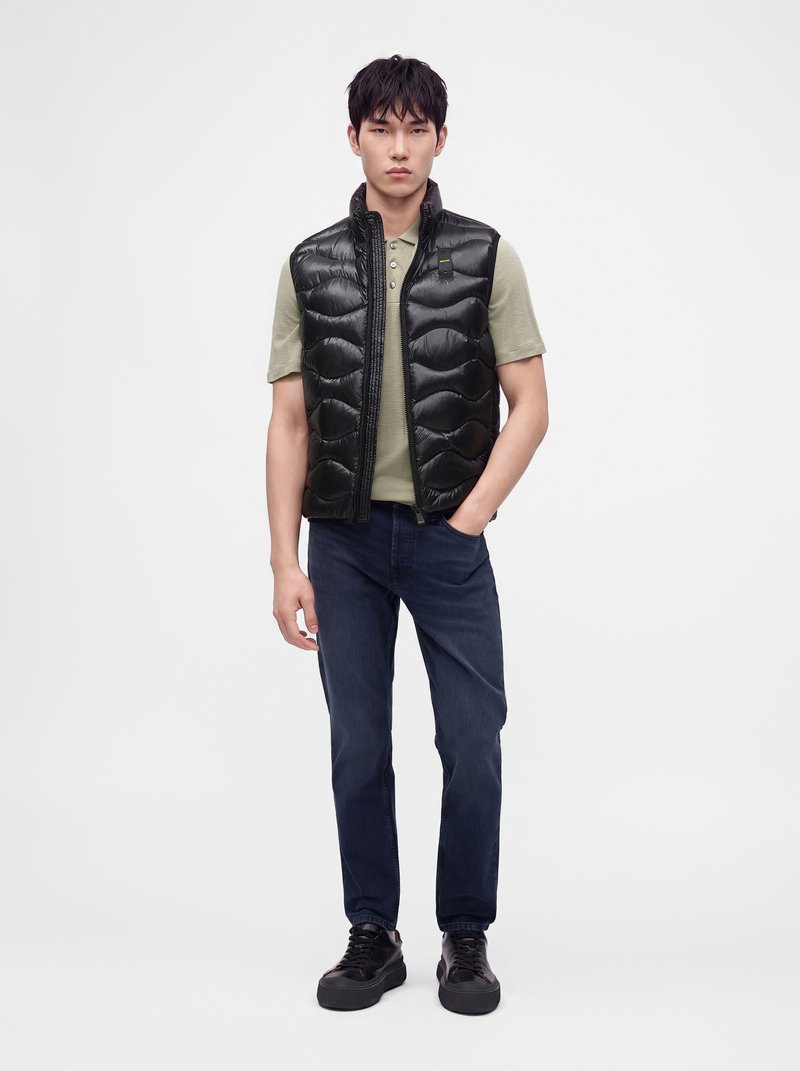 Joven con chaleco acolchado negro, polo verde claro, vaqueros azul oscuro y zapatillas negras, de pie frente a un fondo blanco liso.