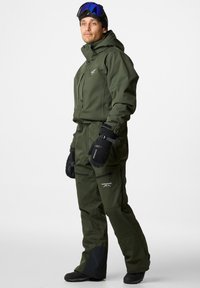 Zielony izolowany kombinezon narciarski z wysokim kołnierzem, wieloma zamykanymi na zamek kieszeniami i regulowanymi mankietami. Czarne akcenty na spodniach i solidne rękawice.