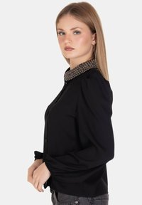 Camicia nera con bottoni, colletto decorato e testurizzato, maniche a sbuffo, realizzata in un tessuto liscio e con una vestibilità sartoriale.