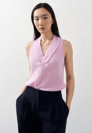 Žena v svetloružovej bezrukáčovej blúzke s výstrihom do V a čiernych vysokým pásom plisovaných nohaviciach, ruky v vreckách, neutrálna mimika tváre, jednoliate pozadie.