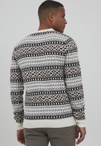 Pull en tricot avec une base blanche ornée de motifs géométriques marron foncé et noirs. Manches et ourlet côtelés, design à col rond.