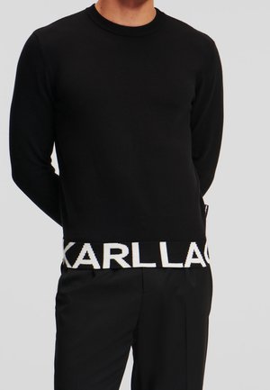 Mand iført en sort langærmet sweater med hvid tekst "KARLLA" delvist synlig ved den nederste kant, kombineret med sorte bukser.