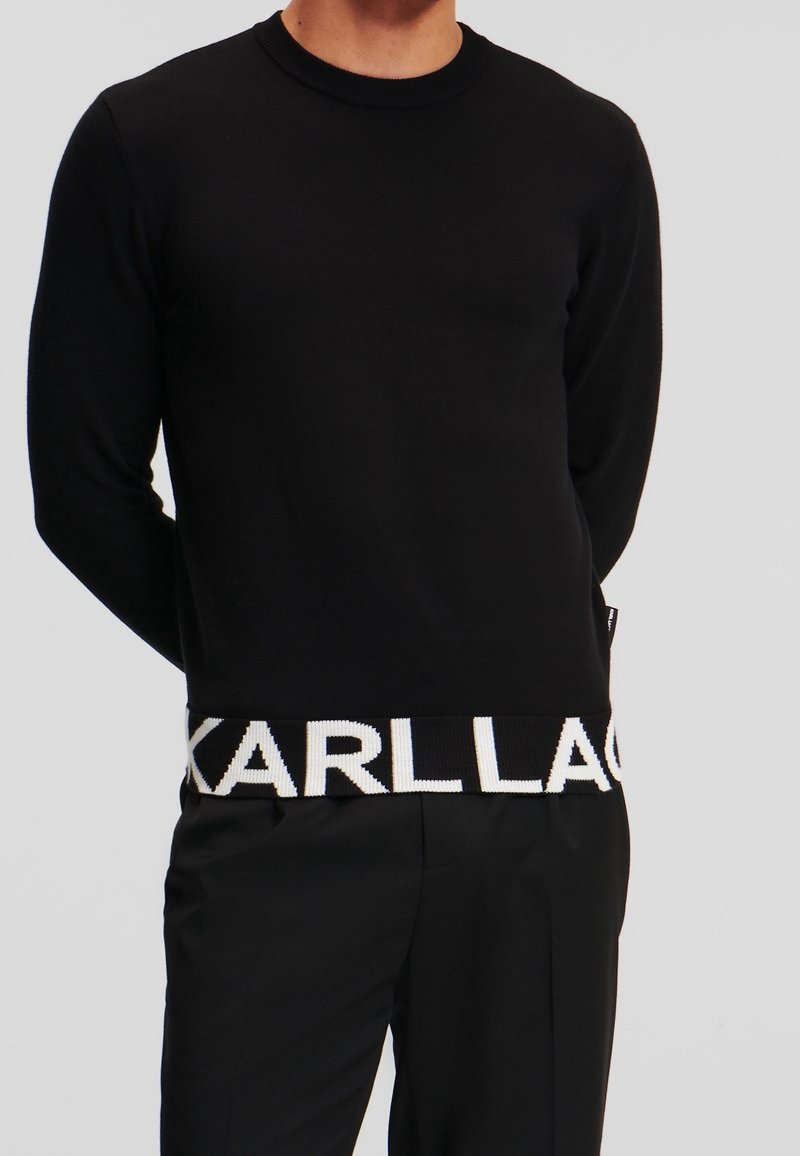 Homme portant un pull à manches longues noir avec le texte blanc "KARLLA" partiellement visible sur l'ourlet inférieur, associé à un pantalon noir.
