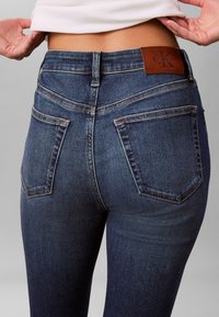 Tamsiai mėlynos džinsinės kelnės su penkiomis kišenėmis, matomomis siūlėmis ir rudu odiniu lopu ant juosmens, kuriame yra stilizuotas „CK“ logotipas.