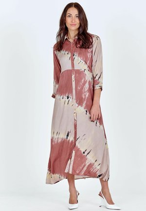 Nő áll hosszú ujjú, gombos tie-dye maxi ruhában, fehér, alacsony sarkú slingback cipőben, sima háttér előtt.