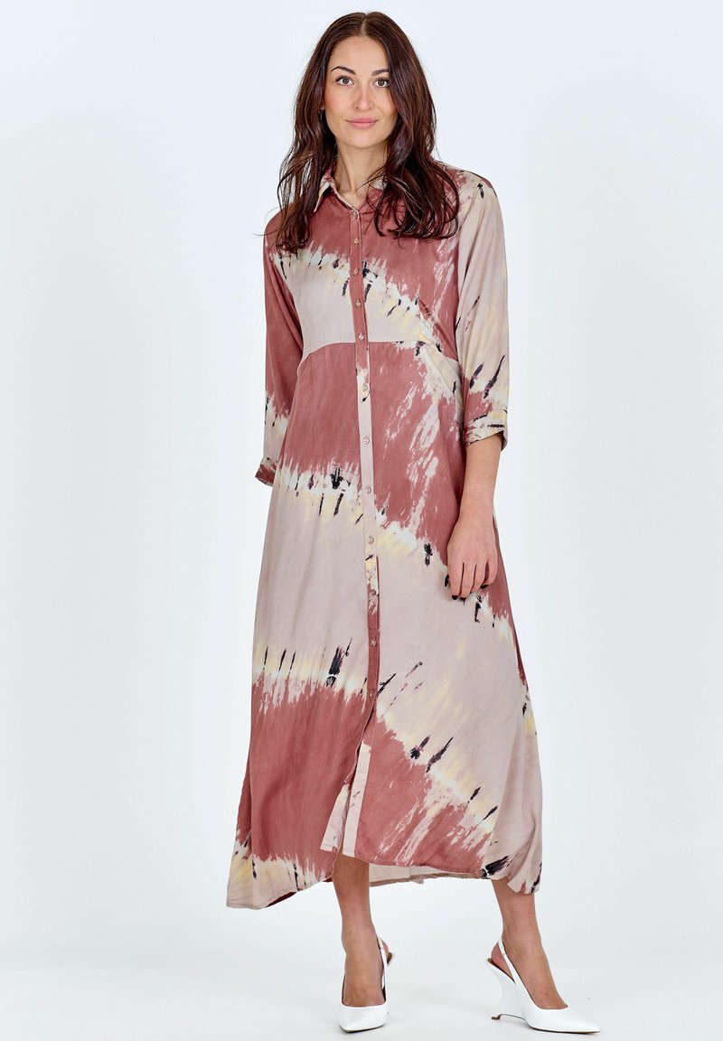 Femme debout dans une robe longue tie and dye à manches longues boutonnée, avec des chaussures slingback blanches à petit talon, sur un fond uni.