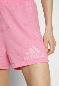 Shorts de sport roses en tissu doux, avec un motif moucheté et un logo Adidas blanc en bas à gauche. Ceinture élastique visible.