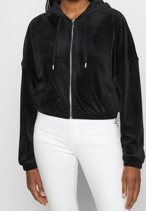 Person, die einen schwarzen Velours-Zip-Hoodie mit Kordeln und weiße Hosen trägt, von den Schultern bis zu den Hüften vor einem schlichten Hintergrund gezeigt.