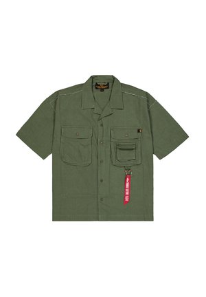 Camisa de manga corta color verde oliva, con botón en la parte delantera, dos grandes bolsillos en el pecho, uno con tapa, y etiqueta de llavero roja con texto. Textura sutil.