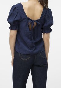 Marinblå topp med puffärmar, texturerad tyg, öppen rygg knuten med bågar, i kombination med mörk denimjeans.