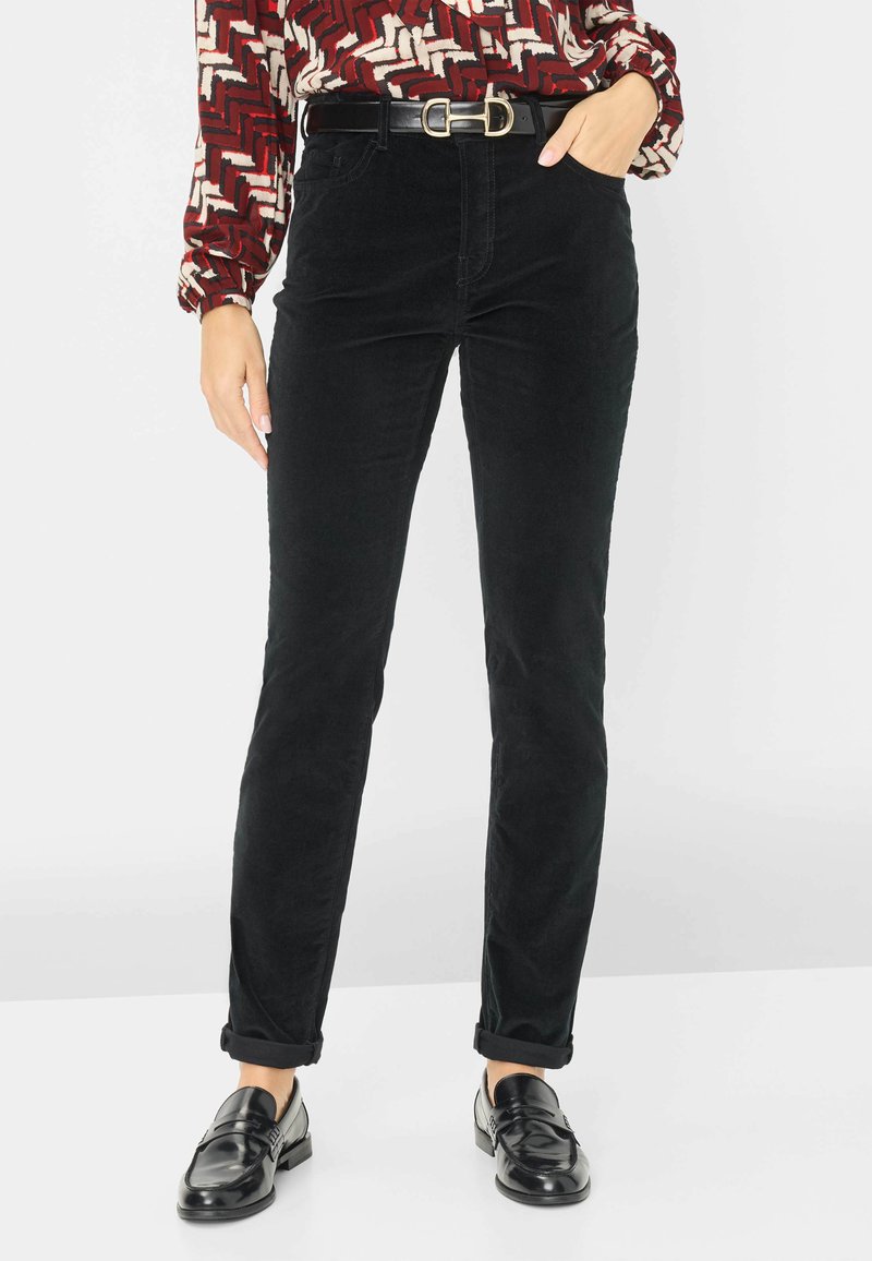 Zwarte corduroybroek met een slim fit, heeft een hoge taille en opgerolde manchetten. Gecombineerd met een gebloemde blouse en zwarte loafers.