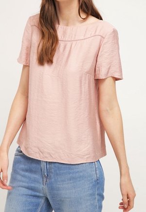 Kobieta w jasnoróżowej bluzce z krótkim rękawem z subtelnymi przeszyciami oraz niebieskich, wysokowładnych dżinsach na neutralnym tle.