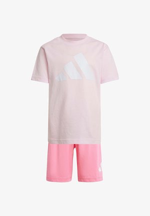 Dos niños sonrientes con camisetas, pantalones cortos y zapatillas Adidas rosas a juego, uno lleva una gorra y el otro lleva una mochila negra.