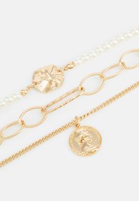 Tre guldtonede armbånd med et struktureret metalled, en række af hvide falske perler og en charm med et cirkulært møntdesign.