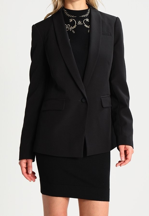 Blazer nero con revers a punta, chiusura a un bottone e due tasche frontali. Indossato sopra un vestito corto nero aderente. Texture liscia.
