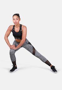 Czarny stanik sportowy z logo na dole; szare legginsy z siateczkowymi panelami; model w pozycji wykroku, noszący czarne buty sportowe.