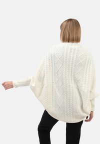 Frau mit kurzem blonden Haar trägt einen weißen Strukturstrickpullover mit Fledermausärmeln und schwarzen Hosen, posiert vor einem hellen Hintergrund.