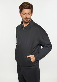 Jachetă bomber ușoară, neagră, cu închidere prin fermoar, guler lat, manșete elastice și buzunare laterale, având o textură netedă și un design minimalist.