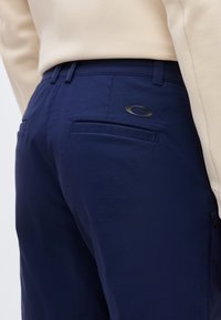 Κοντινό πλάνο ενός ατόμου που φοράει παντελόνι ναυτικού μπλε με πίσω τσέπη και οβάλ μαύρο λογότυπο κοντά στη μέση, συνδυασμένο με μπεζ μπλουζάκι.