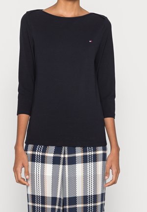 Femme portant un haut noir à manches longues avec un petit logo et un pantalon à carreaux bleu marine, blanc et beige, debout devant un fond uni.