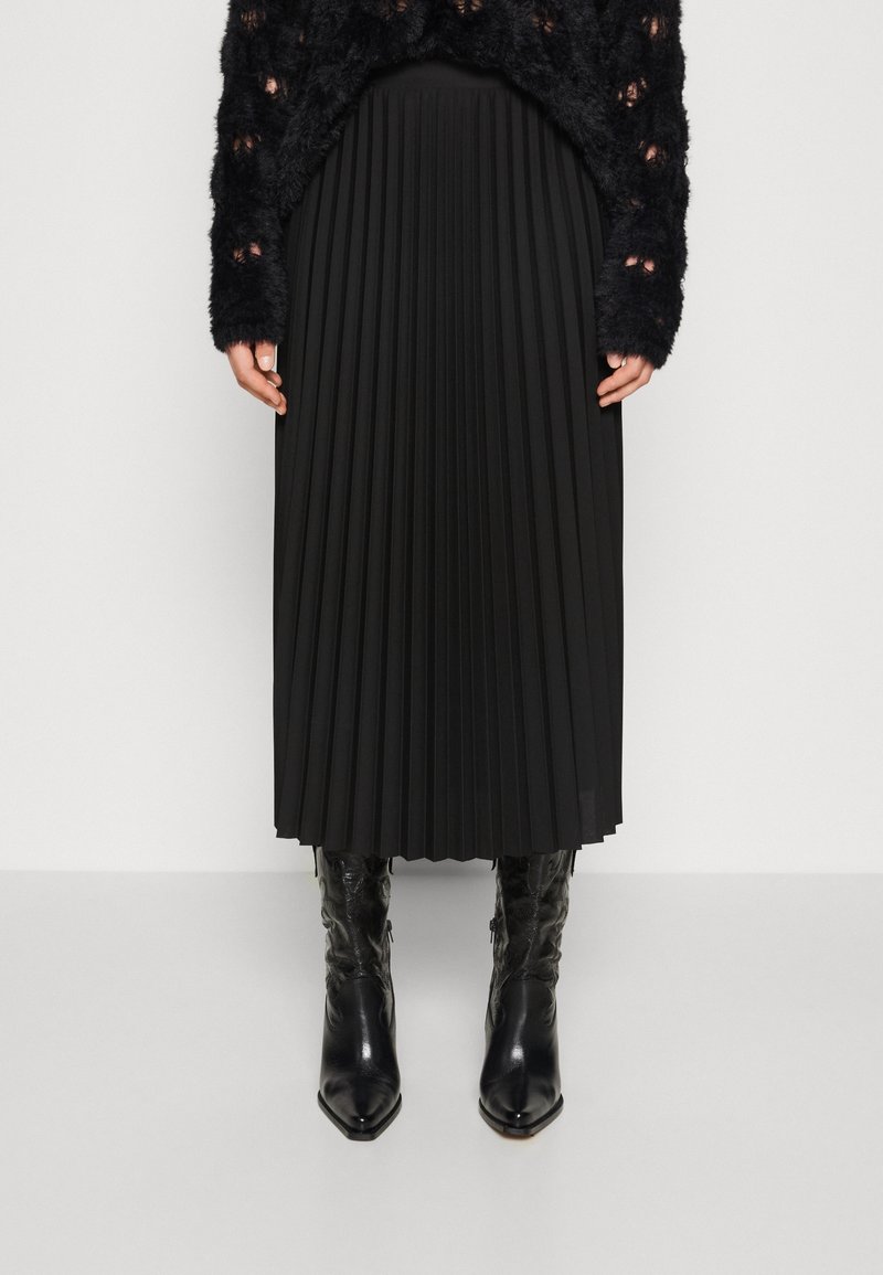 Personne portant un pull noir texturé, une jupe midi plissée noire et des bottes en cuir noires à bout pointu montant jusqu'au milieu du mollet, debout devant un fond uni.