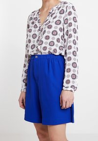 Langärmlige weiße Bluse mit einem geometrischen roten und blauen Muster, kombiniert mit knielangen blauen Shorts mit einem elastischen Bund.