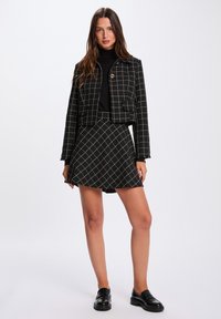 Giacca a quadri in bianco e nero corta con bottone dorato, abbinata a una gonna A-line coordinata. La modella indossa un dolcevita nero e mocassini.