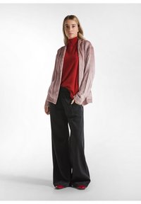 Lange zwarte wijde broek, rode ribbeltrui en een lichtroze geweven jas die open wordt gedragen. Model staat casual met een hand in de zak.