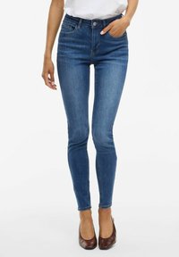 Jeans skinny en denim bleu avec taille haute, design cinq poches et une texture légèrement délavée. Assortis avec des chaussures à talons marron.