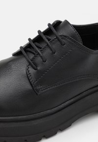 Scarpa in pelle nera con finitura liscia, dotata di cinque occhielli, lacci piatti e una suola in gomma spessa con un motivo di battistrada testurizzato.